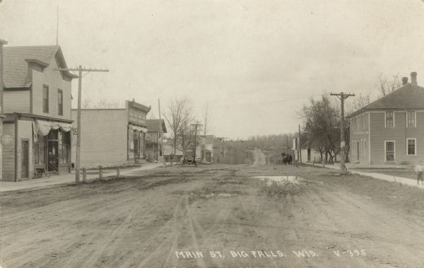 Photographic postcard view down center of unpaved street in the central business district of a rural community. On the left is a general store, and on the right the Big Falls House boarding house. A car parked near the sidewalk down the street on the left, and a horse and buggy further down on the right. There is a pedestrian on the sidewalk on the right. Caption reads: "Main St. Big Falls, Wis."