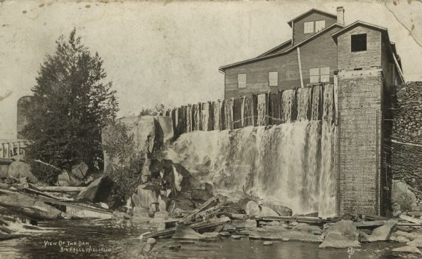 Photographic postcard view across a stream of a dam and adjoining building. There appears to be a silo behind the tree on the left.