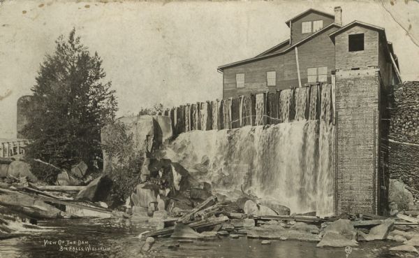 Photographic postcard view across a stream toward a dam and adjoining building. There appears to be a silo behind the tree on the left. Caption reads: "View of the Dam, Big Falls, Wisconsin."