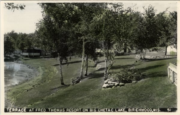 Colorized photographic postcard view of a lawn with stone terraces and birch trees leading down to a lake on the left. There is a cabin in the background near the shore of the lake, and other buildings in the center and on the right. Caption reads: "Terrace at Fred Thomas Resort on Big Chetac Lake, Birchwood, Wis."