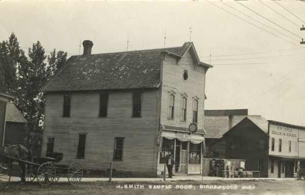 Photographic postcard view across street towards the Herman Smith Saloon storefront, with a man and two dogs on the sidewalk in front. A horse and buggy are parked on the far left. Crates and barrels are stacked along a fence to the right of the saloon. Further down the street is the M.P. Towle horse dealership. Caption reads: "H. Smith Sample Room, Birnamwood, Wis."