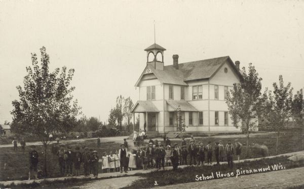 Slightly elevated view of a school with the students gathered on the board sidewalk. Adults are gathered near the front steps of the entrance to the school. Caption reads: "School House, Birnamwood, Wis."
