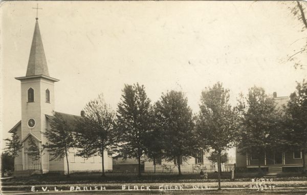 Photographic postcard view of a town block with a church and two houses along a tree-lined street. Two young children are standing on the sidewalk.