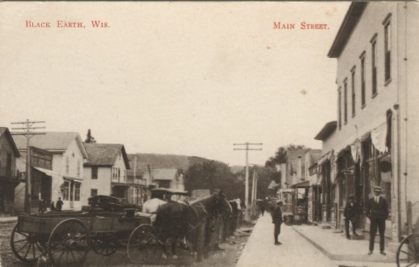 Photographic postcard view down sidewalk in a central business district. In the foreground on the left horses and wagons are hitched up at the curb. A small group of people are gathered in front of the storefront on the right. Caption reads: "Black Earth, Wis. Main Street."