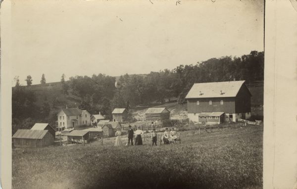 Photographic postcard view of a family posed on a hill in front of a farm, with farmhouse, outbuildings, and a windmill. The family is gathered on a hill in the foreground with a dog. Two people sit in a cart pulled by sheep.