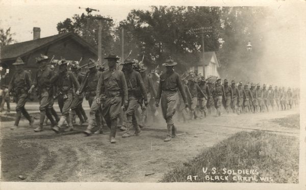 Photographic postcard view of troops marching on the road across the railroad tracks past the depot.
