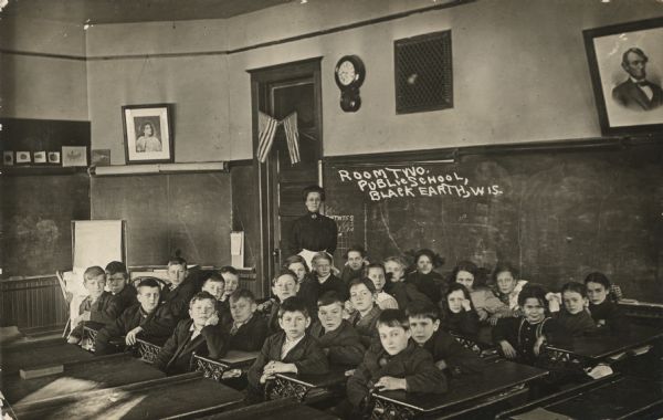 Students are posing sitting sideways at their desks in the classroom for a group portrait. The teacher is standing in the back near a door.