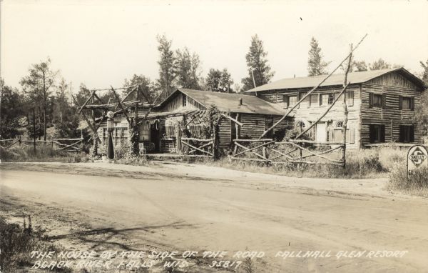 Photographic postcard view of a log resort and filling station on the side of a dirt road. Caption reads: "The House by the Side of the Road — Fallhall Glen Resort, Black River Falls, Wis."