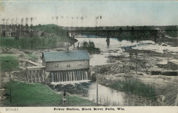 Colorized photographic postcard of an elevated view of the power station next to the river. A bridge is in the background. Caption reads: "Power Station, Black River Falls, Wis."