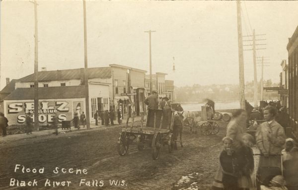 Photographic postcard view of a street scene. Groups of people are gathered along the sidewalks near storefronts. A building on the left has a painted sign for "Selz Royal Blue Shoes" painted on its side, and men stand near a barber pole on the sidewalk in front. Further down the block is the City Bakery. In the foreground on the right two women stand on the sidewalk with children and a baby in a baby carriage. In the background is the river and the far shoreline with houses on a hill. Caption reads: "Flood Scene, Black River Falls, Wis."