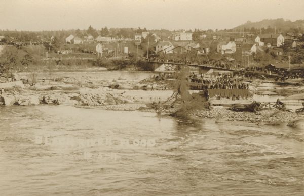 Photographic postcard view of the aftermath of a flood. Crowds are gathered on the river bank, the bridge and the higher ground in the background. Caption reads: "Black River Flood."