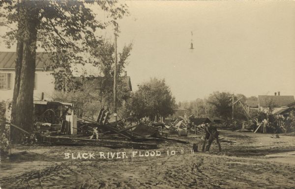 Photographic postcard view of the aftermath of a flood in a residential neighborhood. A man is picking up pieces of wood in the road. Large piles of debris are piled up against tree and buildings. Caption reads: "Black River Flood."