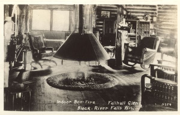 An interior view of a room with a fire pit in the center surrounded by rocking chairs. Caption reads: "Indoor Bonfire — Fallhall Glen, Black River Falls, Wis."