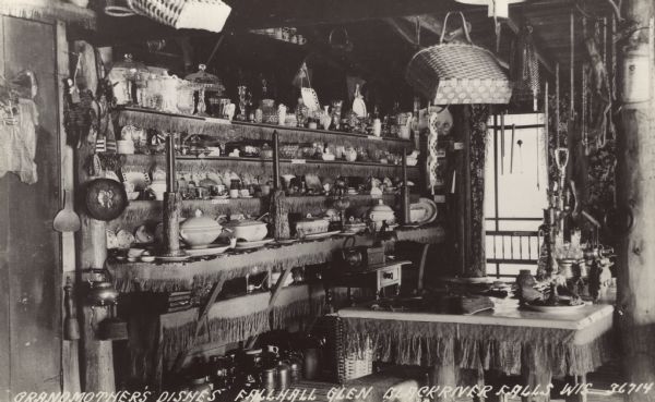 Interior view of shelves and tables with dishes and serving dishes. There are baskets hanging from the ceiling. Caption reads: "Grandmother's Dishes — Fallhall Glen, Black River Falls, Wis."