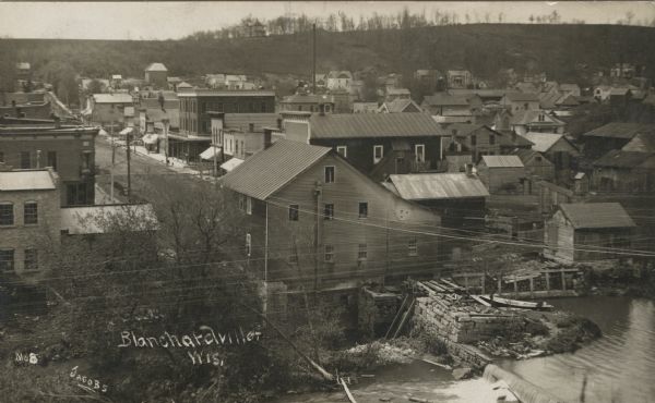 Elevated photographic view of the town of Blanchardville with the commercial district on the left, and residential neighborhoods on the right. The river is in the foreground. Caption reads: "Blanchardville, Wis."