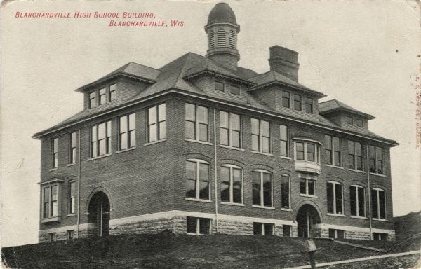 Exterior view of a large brick high school with a belfry and large glass windows. Caption reads: "Blanchardville High School Building, Blanchardville, Wis."