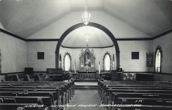Photographic postcard view of the interior of the Lutheran Church. Pews and hymnals are on each side, and flags are at the altar. A piano is near the pulpit. Caption reads: "Interior — Lutheran Church, Blanchardville, Wis."