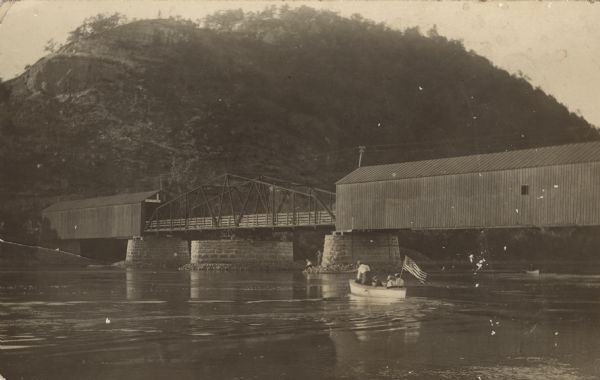 Photographic postcard view of a covered bridge over the Wisconsin River. Men are fishing at the base of a pylon, and other men are boating in the river. There is a bluff in the background.