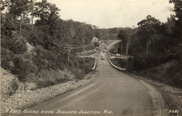Photographic postcard view downhill towards an intersection in the north woods. Three cars are driving away up the hill on the other side of the intersection. Caption reads: "A Road Scene near Boulder Junction, Wis."