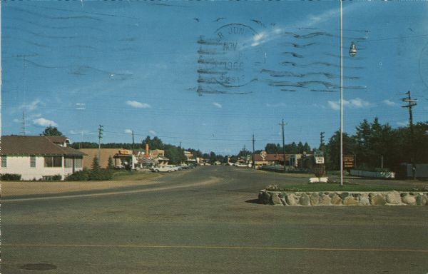 View of the commercial district. Cars are parked in front of businesses. A flagpole and a stop sign are on a highway island in the foreground.