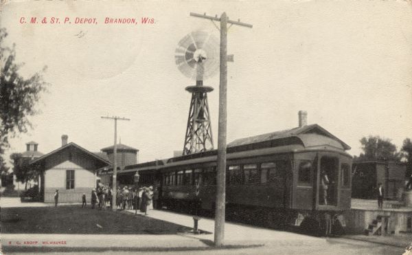 Photographic postcard view of the depot and train with passengers on the platform. A large windmill is in the background. Caption reads: "C. M. & St. P. Depot, Brandon, Wis."