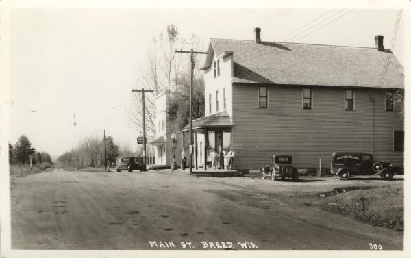 Photographic postcard view down the street towards the central business district; a post office, a gas pump and a bar. Two cars, a truck and some people are outside the post office. Caption reads: "Main St., Breed, Wis."