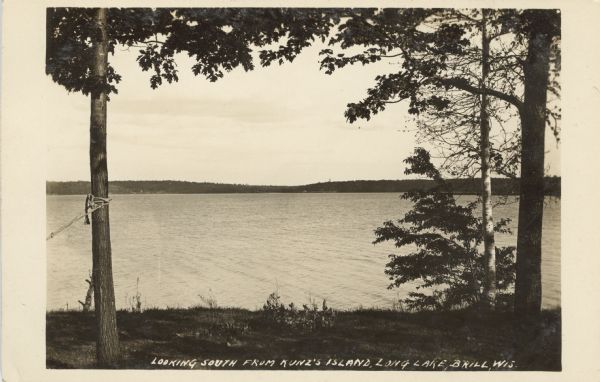 Photographic postcard view of a lake from an island. Dwellings are on the far shoreline among trees. Caption reads: "Looking South from Kunz's Island, Brill, Wis."
