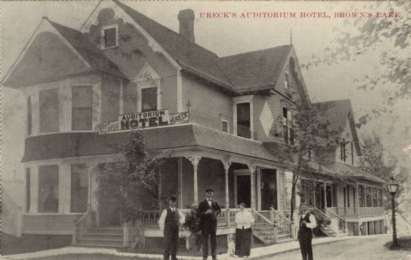 View of the exterior of a hotel with a porch. People on posing on the sidewalk in front. Caption reads: "Ureck's Auditorium Hotel, Brown's Lake."
