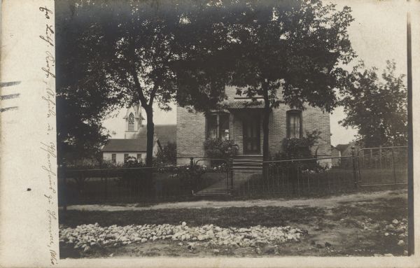 Photographic postcard view of a brick house with a garden in the gated front yard. A church is in the background on the left.