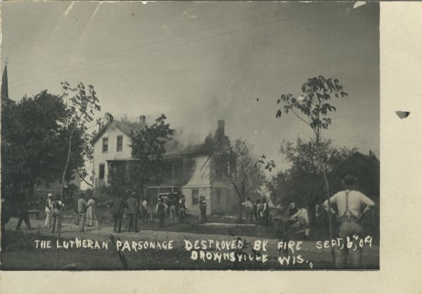 Photographic postcard view of the aftermath of a fire. Most of the parsonage is destroyed. There are onlookers on the lawn in front of the house. Caption reads: "The Lutheran Parsonage Destroyed By Fire Sept. 6, '09, Brownsville, Wis."