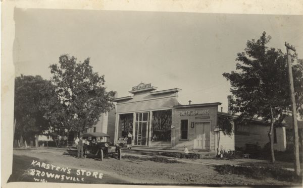 Photographic postcard view across street towards the exterior of a general store. A woman is standing by the door, and a boy on a wagon is on the sidewalk. An automobile is parked at the curb. Caption reads: "Karsten's Store, Brownsville, Wis."