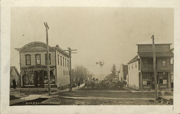 Photographic postcard view of an intersection with commercial buildings on both corners. Several horses and carts are on the street, and a windmill is in the background. Caption reads: "Scene at Browntown, Wisconsin."