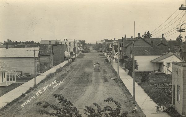 Elevated photographic postcard view of Main Street with horses and carts along the street. Dwellings and commercial buildings are on both sides of the street, including a drugstore and a harness shop. Caption reads: "Main Street, Bruce, Wis."
