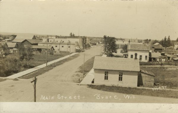 Elevated photographic postcard view of Main Street. An unused lot is in the left foreground. The rest of the street is lined with dwellings and commercial buildings. Caption reads: "Main Street, Bruce, Wis."