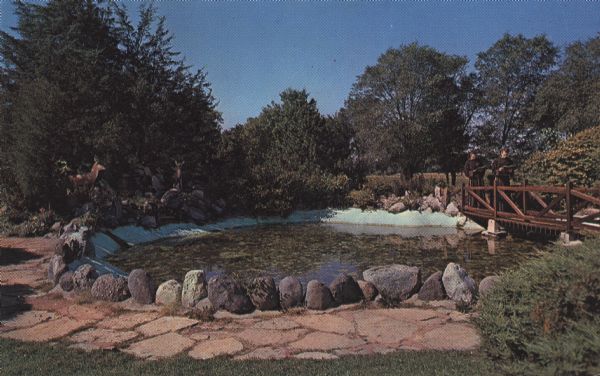 Color postcard view of a rock garden with a rustic bridge and koi pond. Two monks are reading on the bridge.