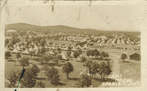 Elevated view of Fort McCoy with rows of tents among trees. There is a row of automobiles parked in the center, and a row of barracks on the left. Caption reads: "Camp McCoy, Sparta, Wis."