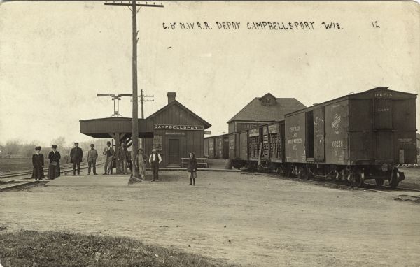 View of a depot and freight cars. A group of passengers is standing in a row in front. Caption reads: "C. & N.W.R.R. Depot, Campbellsport, Wis."