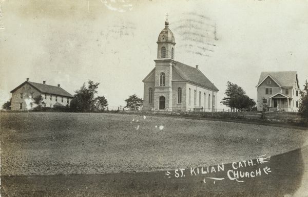 View of a church and neighboring dwellings. A field is in the foreground. Caption reads: "St. Kilian Cath. Church."