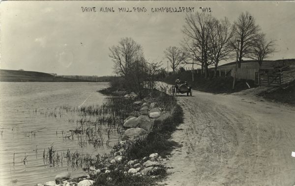 View of an automobile driving down a road bordering a pond on the left. On the right is fencing and a wooden gate and a hill. Caption reads: "Driving Along Mill Pond, Campbellsport, Wis."