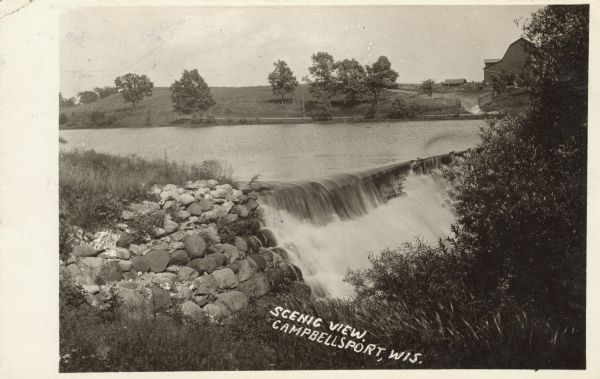 View of a river with a dam waterfall. A road runs along the far side of the river, and a large barn is across the road on the right. Caption reads: "Scenic View, Campbellsport, Wis."