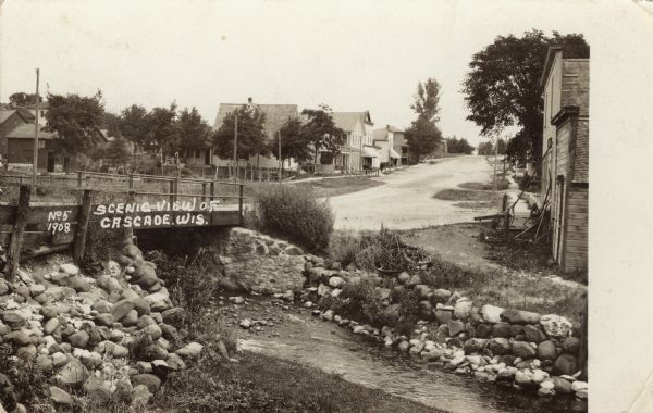 View of a street going through the center of town. In the foreground is a stream and a bridge. Commercial buildings and dwellings are on the left.