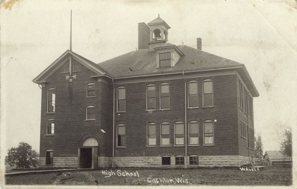View of exterior of the high school, a brick building with a belfry in the center of the roof, and a flagpole going through a gable in the roof on the left front. Caption reads: "High School, Cashton, Wis."
