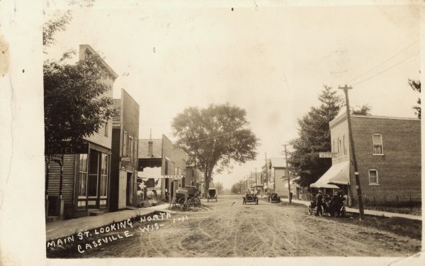 Photographic postcard view looking down Main Street. A hotel and livery are on the left, with another hotel and jewelry store on the right. Automobiles and carts are parked in the street. Caption reads: "Main St. Looking North, Cassville, Wis."
