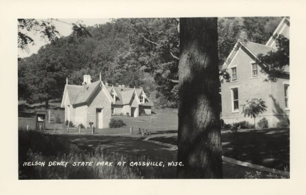 Photographic postcard view of buildings at the Nelson Dewey State Park, including the smokehouse. A tree-covered bluff is in the background. Caption reads: "Nelson Dewey State Park at Cassville, Wisc."