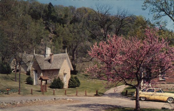 Color postcard featuring the redbud tree at Stonefield Village. The smokehouse is on the left. An automobile is parked in the drive.