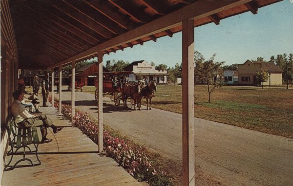Color postcard of Main Street from porch on building at Stonefield Village. A horse bus with tourists is in the street.