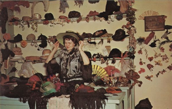 Color postcard of the Millinery Shop at Stonefield. A woman is standing at the counter putting on a hat. Hats are displayed on the shelves behind her.
