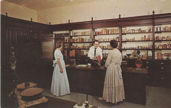 Color postcard of the interior of the pharmacy, with a male druggist and two female customers.