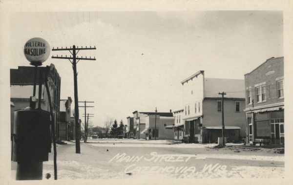 Photographic postcard view of a Main Street in winter, with a gas pump in the lower left and commercial buildings lining the street. Caption reads: "Main Street — Cazenovia, Wis."