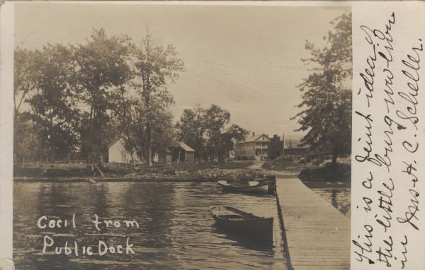 Photographic postcard from the end of the public dock looking inland towards buildings on the shoreline. Caption reads: "Cecil from Public Dock."